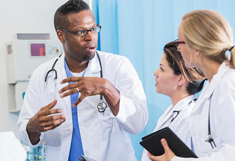 A Black clinician in a white coat speaks to two female medical residents.