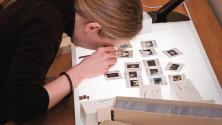 A woman uses a magnifying glass to view slides of medical images on a light table